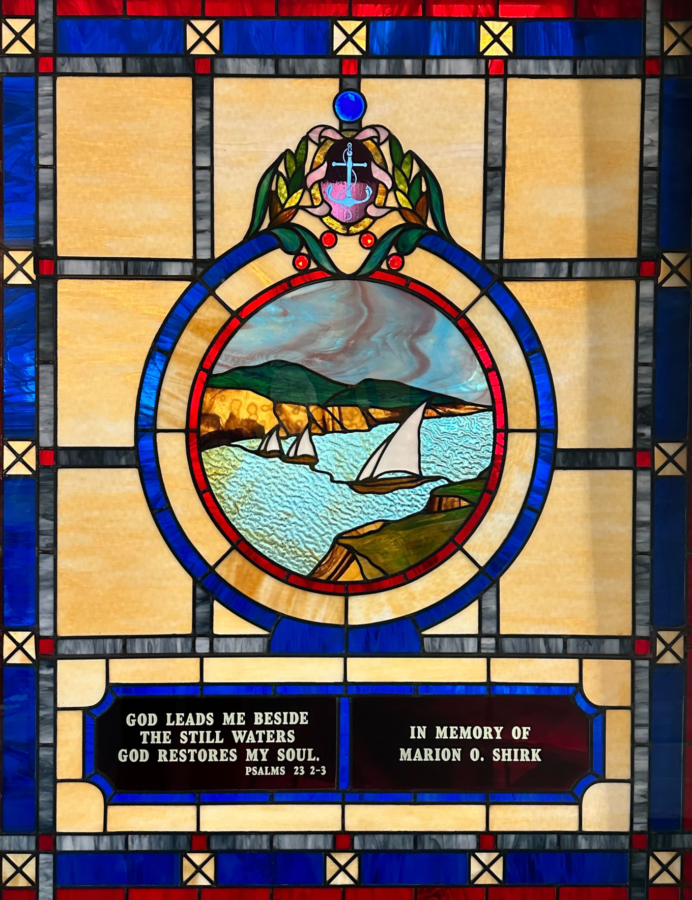 Stained glass window with ships crossing a river between green cliffs. It reads "God Leads Me Beside The Still Waters God Restores My Soul. (-Psalms 23 2-3) In Memory of Marion O. Shirk